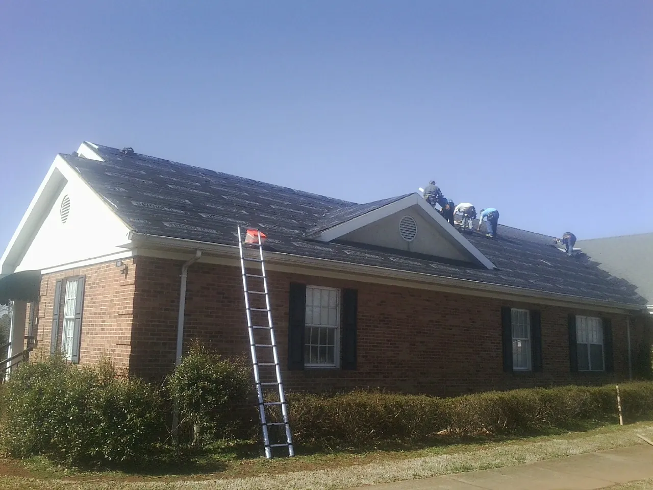 Crew tearing shingles off a brick ranch roof with a ladder set — Emergency Roof Repair work by our crew in Yaphank