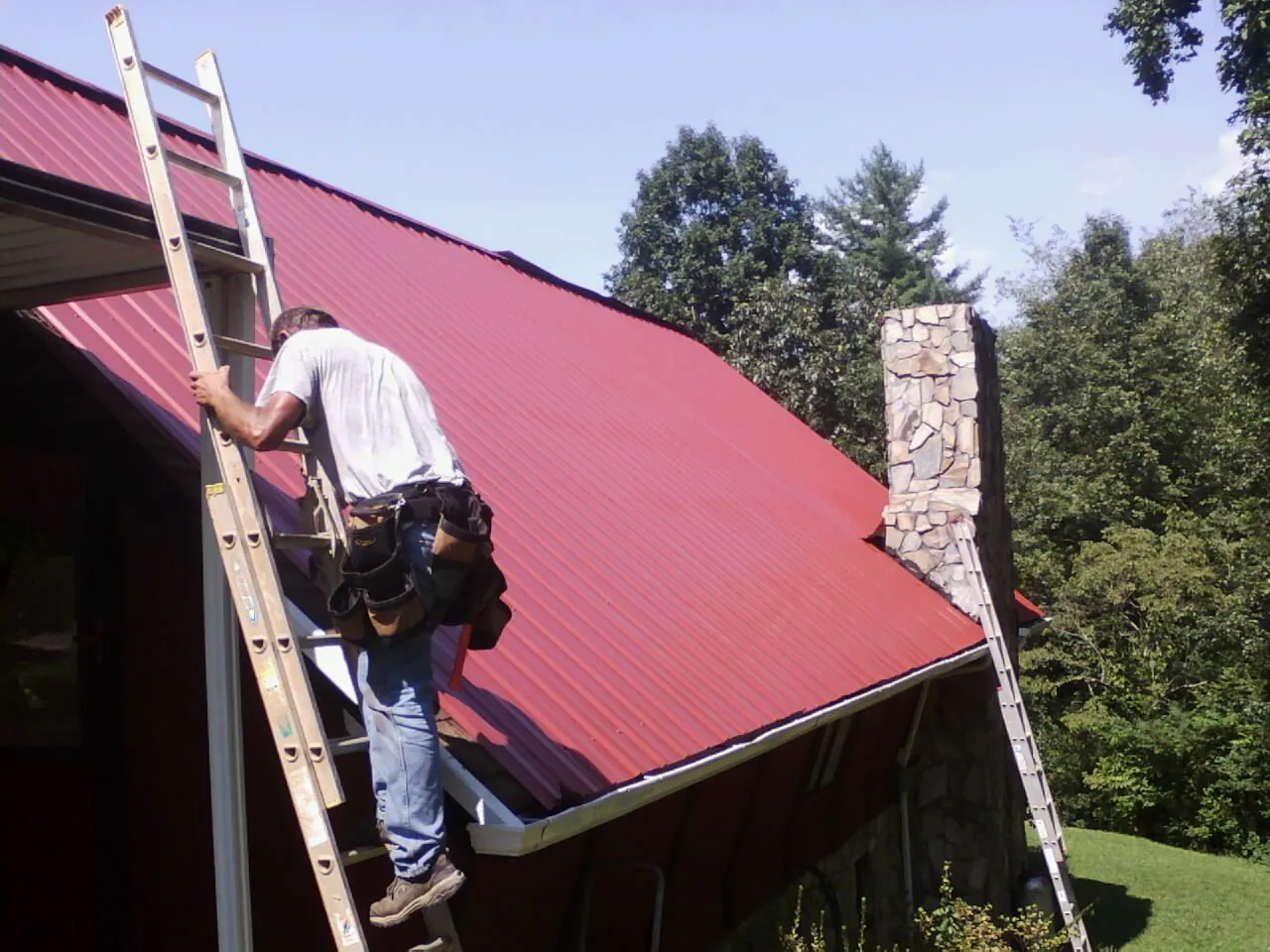 Roofer on a ladder beside a red metal roof and stone chimney — Soffit & Fascia Repair by Roofing Brothers in Yaphank