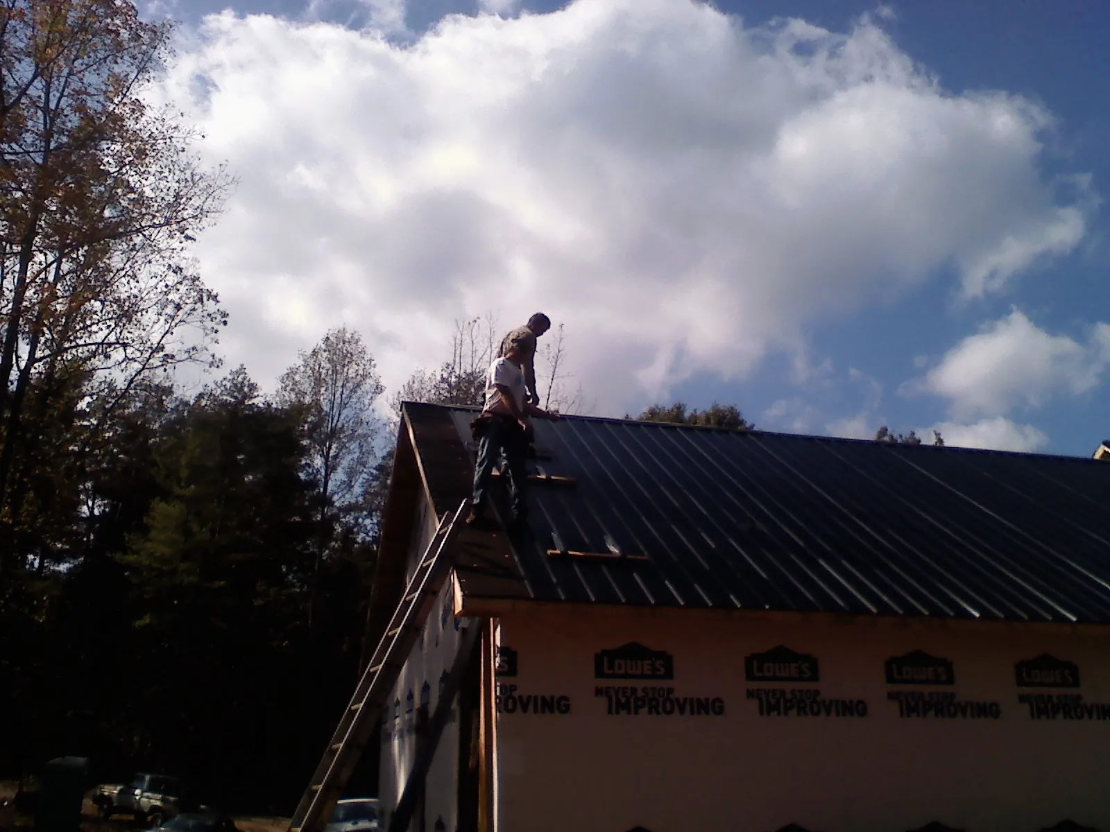 Roofer fastening dark metal panels high against a cloudy sky, from a Roof Inspection job in Yaphank